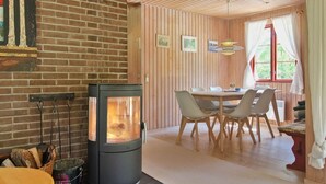 A cozy room with a brick wall, a wood-burning stove, a dining table with chairs, and a window with curtains. - "Chenoa" - 200m from the sea by Interhome (Aakirkeby)