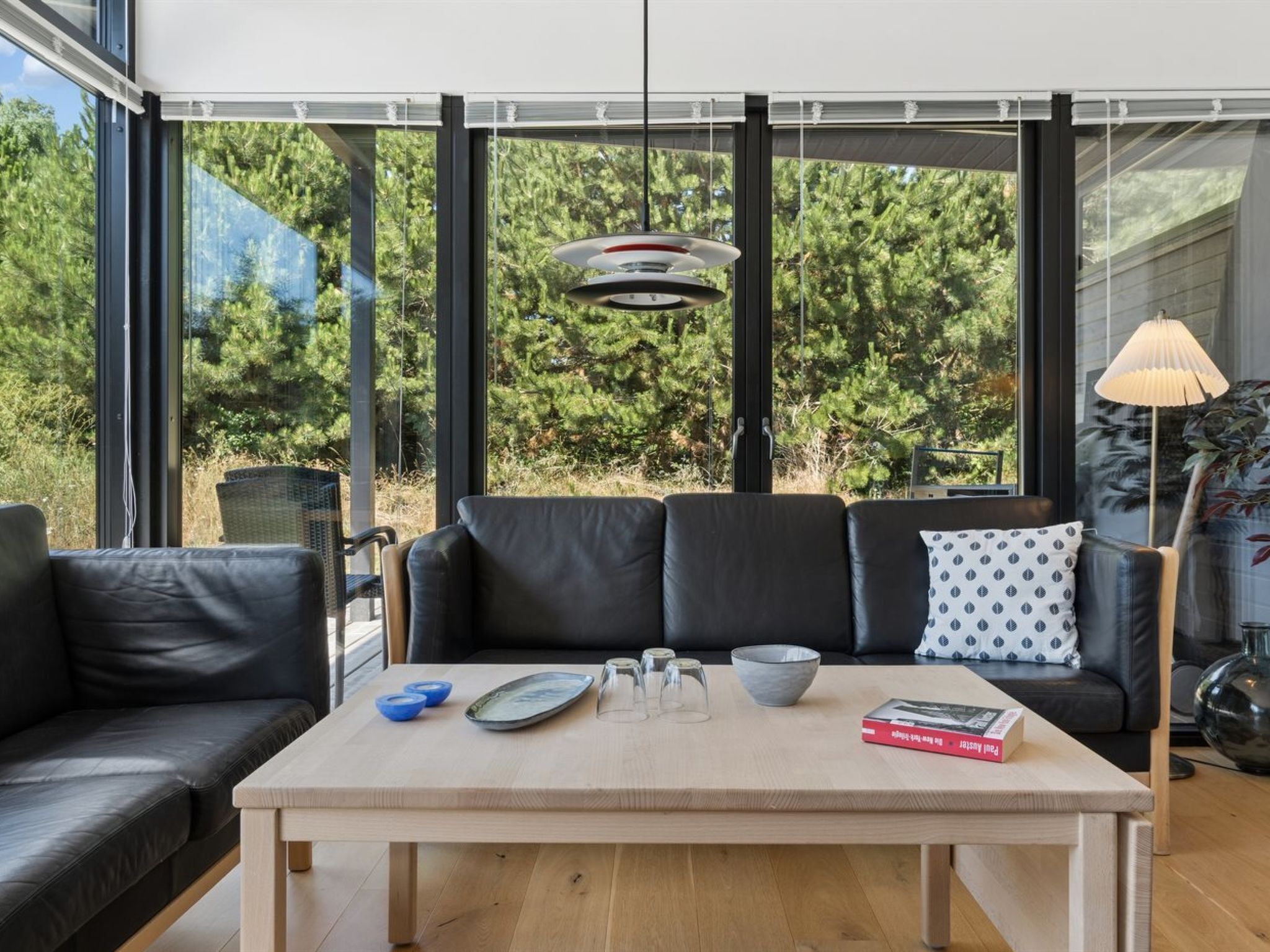 A modern living room with a black leather sofa, a wooden coffee table, and a large window with a view of trees.