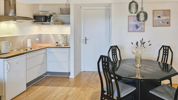 Een compacte keuken met een magnetron, broodrooster en spoelbak. Er is een eettafel met stoelen en een vaas met bloemen.