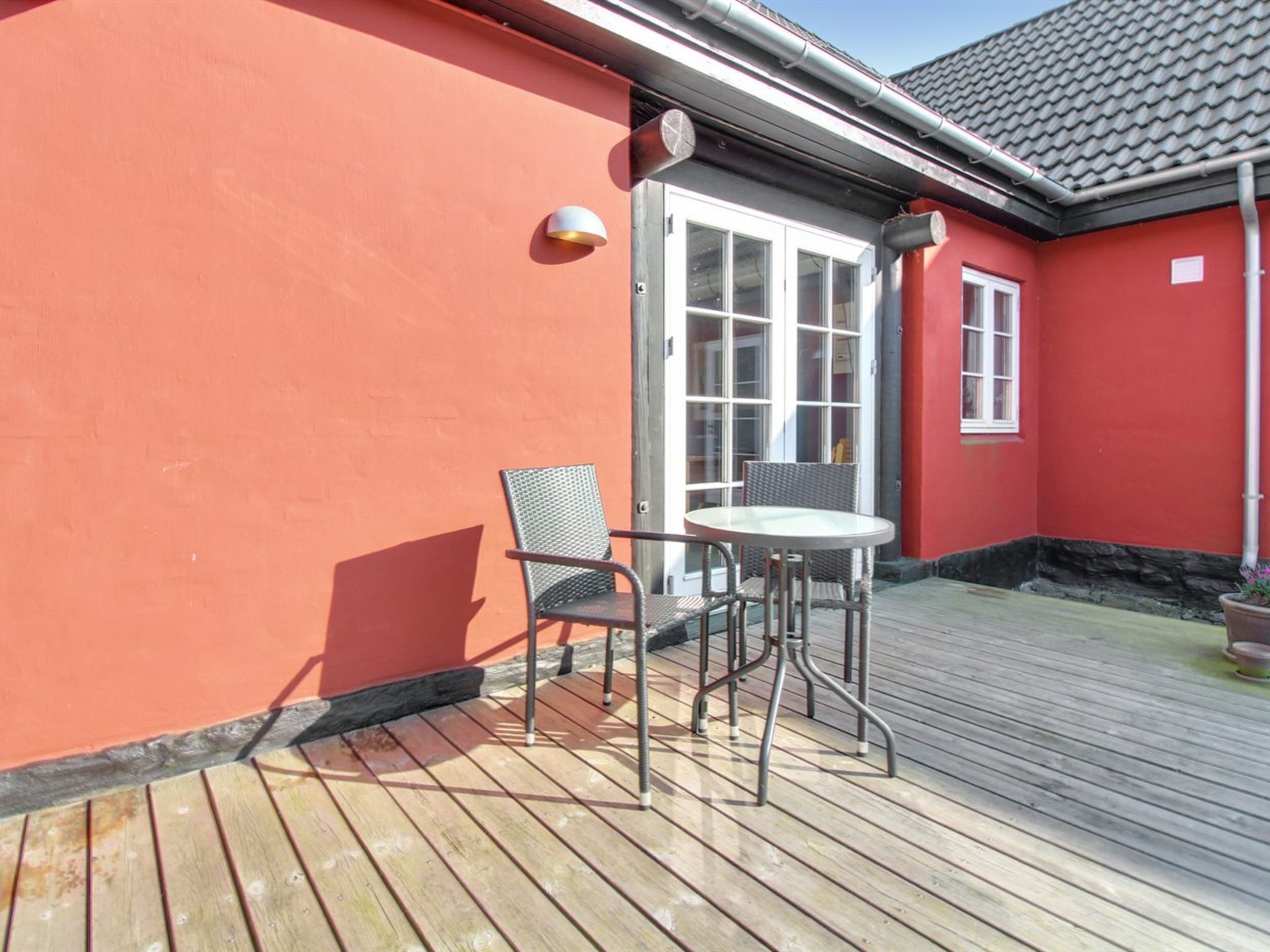 Une terrasse en bois avec une table et deux chaises, un mur rouge et une fenêtre aux cadres blancs.