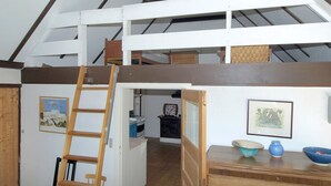 Un espacio tipo loft con una escalera de madera que lleva a una zona de cama elevada, una cocina con estufa y una zona de comedor con mesa y sillas.