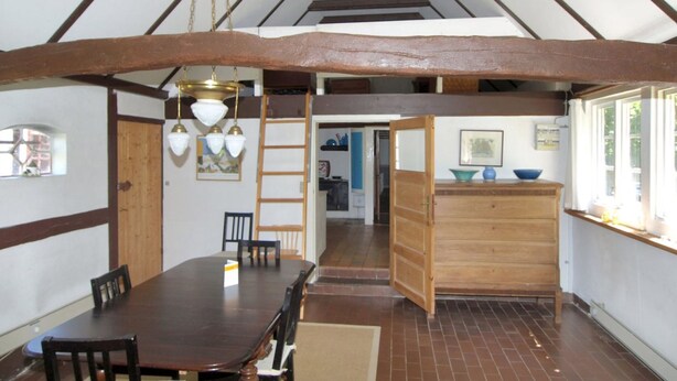Un comedor con una mesa y sillas de madera, una lámpara colgante y vista a una cocina a través de un puerta abierta.