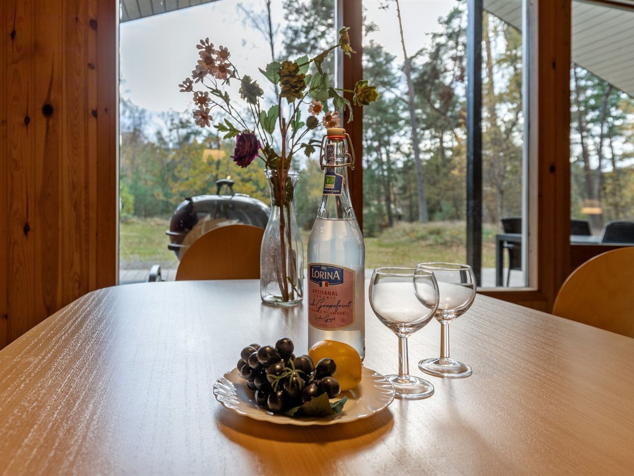 A wooden table with a bottle of liquor, two wine glasses, a plate with grapes and an orange, and a vase with flowers.