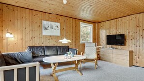A cozy room with a wooden ceiling, a black leather sofa, a white table, a TV, and a window.