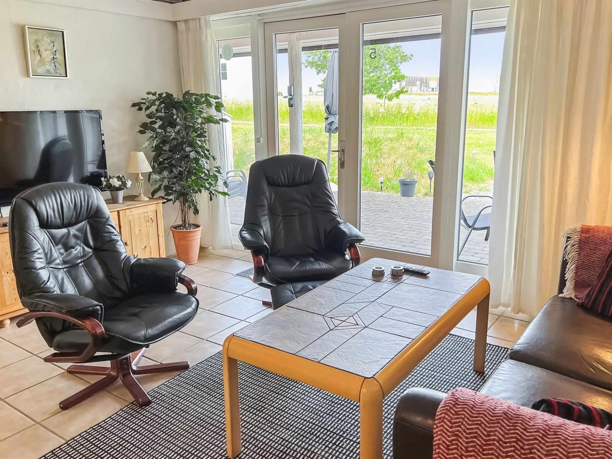 Een moderne woonkamer met twee lederen fauteuils, een houten salontafel en een bank. Links staat een televisie en bij het grote raam is er uitzicht op een groen landschap.