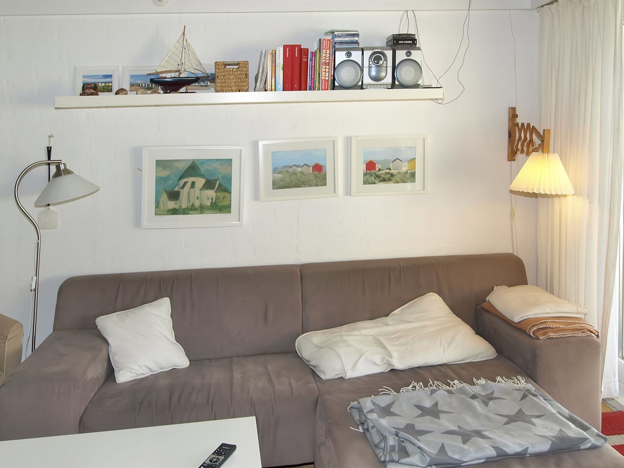 Una sala de estar con un sofá marrón, una mesa de centro blanca y una lámpara. Hay cuadros enmarcados en la pared, una estantería con libros y un altavoz, y una ventana con cortinas.