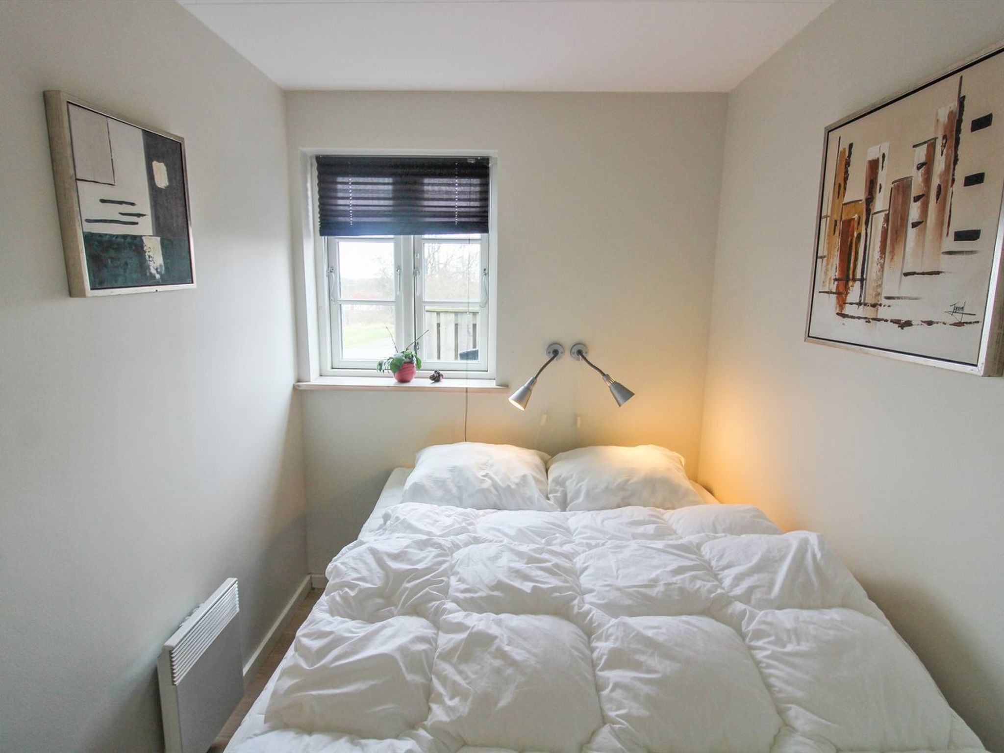 Een slaapkamer met een bed, een raam met zonwering, een radiator en twee wandlampen.