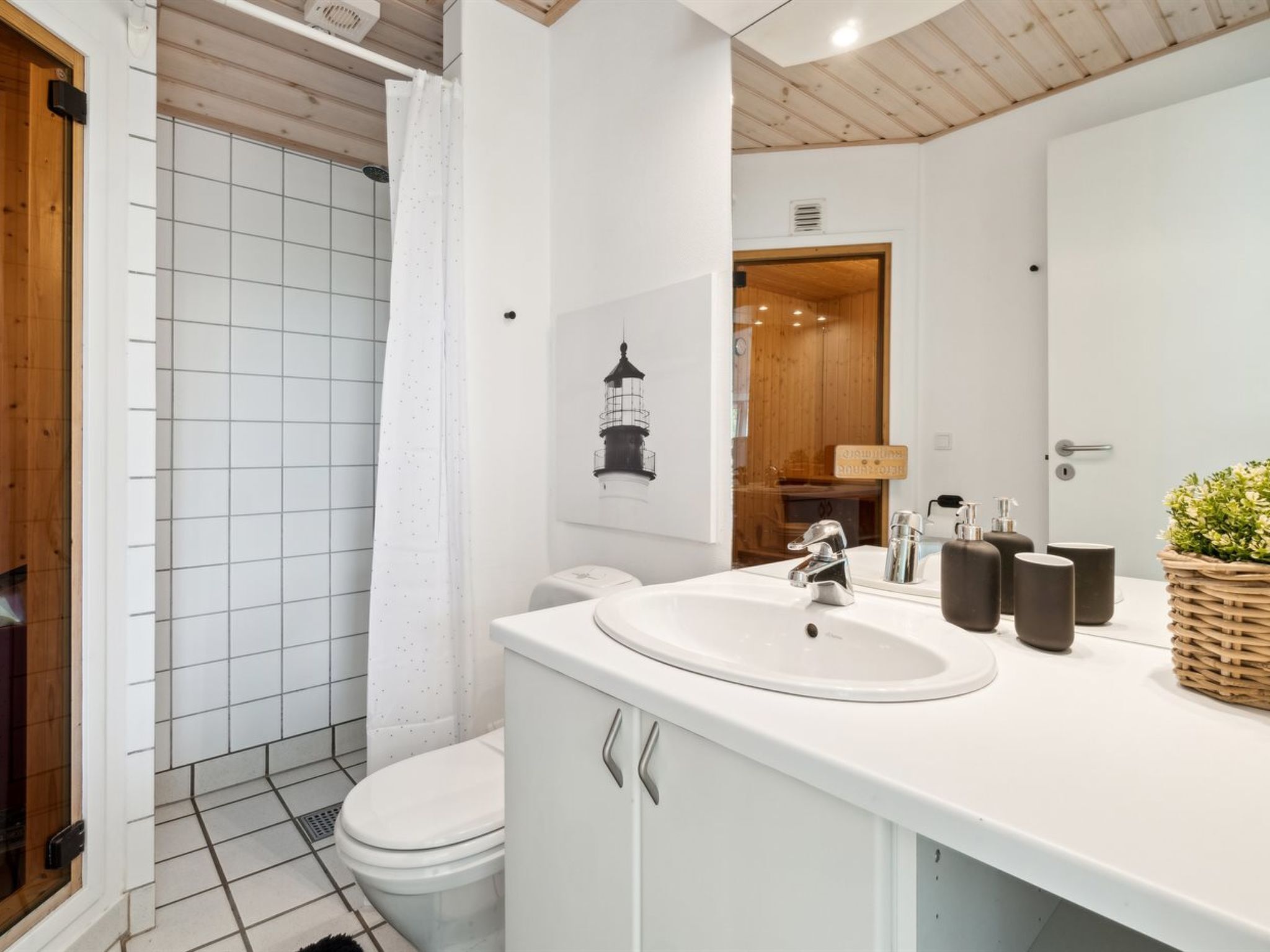 A modern bathroom with a white sink, toilet, and shower. There is a wooden door and a mirror above the sink.