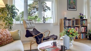 Ein gemütliches Wohnzimmer mit einer Couch, einem Sessel, einem Couchtisch mit Büchern und Tassen, einem Bücherregal und einem Fenster mit Blick auf Bäume.