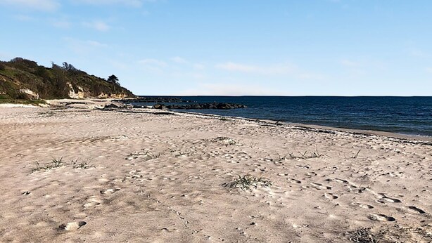 Ein sandiger Strand mit Fußspuren und vereinzelter Vegetation, ein Felsvorsprung und ein klarer, blauer Himmel.