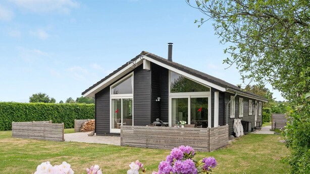 Une maison moderne à deux étages, dotée d’une grande façade vitrée, d’un patio couvert et d’un jardin bien entretenu agrémenté d’arbustes à fleurs.