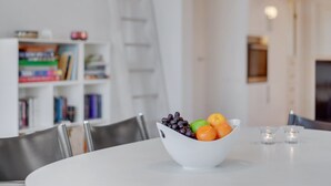 Un coin repas avec une table blanche, un saladier de fruits et une bibliothèque en arrière-plan.