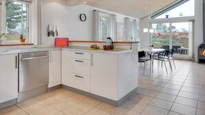 Une cuisine moderne avec des meubles blancs, un lave-vaisselle intégré et un grand îlot central doté d’un évier. On y trouve également un coin repas avec une table et des chaises, ainsi qu’une cheminée en arrière-plan.
