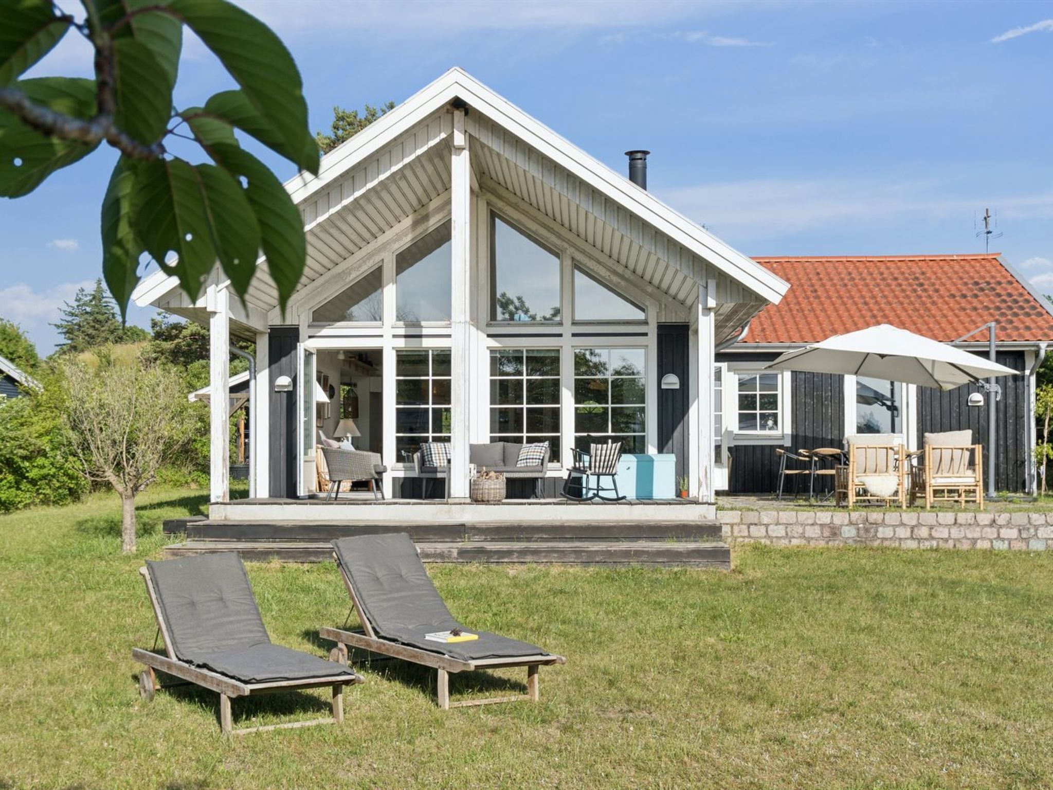 Et moderne hus med stor glasfacade, en overdækket terrasse med udendørs møbler og to liggestole i haven.