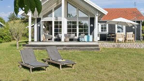 Une maison moderne avec une grande façade vitrée, un patio couvert avec des meubles d’extérieur et deux fauteuils de jardin.