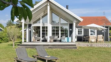 Et moderne hus med stor glasfacade, en overdækket terrasse med udendørs møbler og to liggestole i haven.