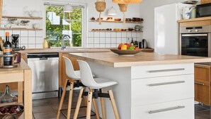 Une cuisine moderne dotée d’un grand îlot central, de meubles blancs, de luminaires suspendus en osier et offrant une vue sur la verdure environnante.