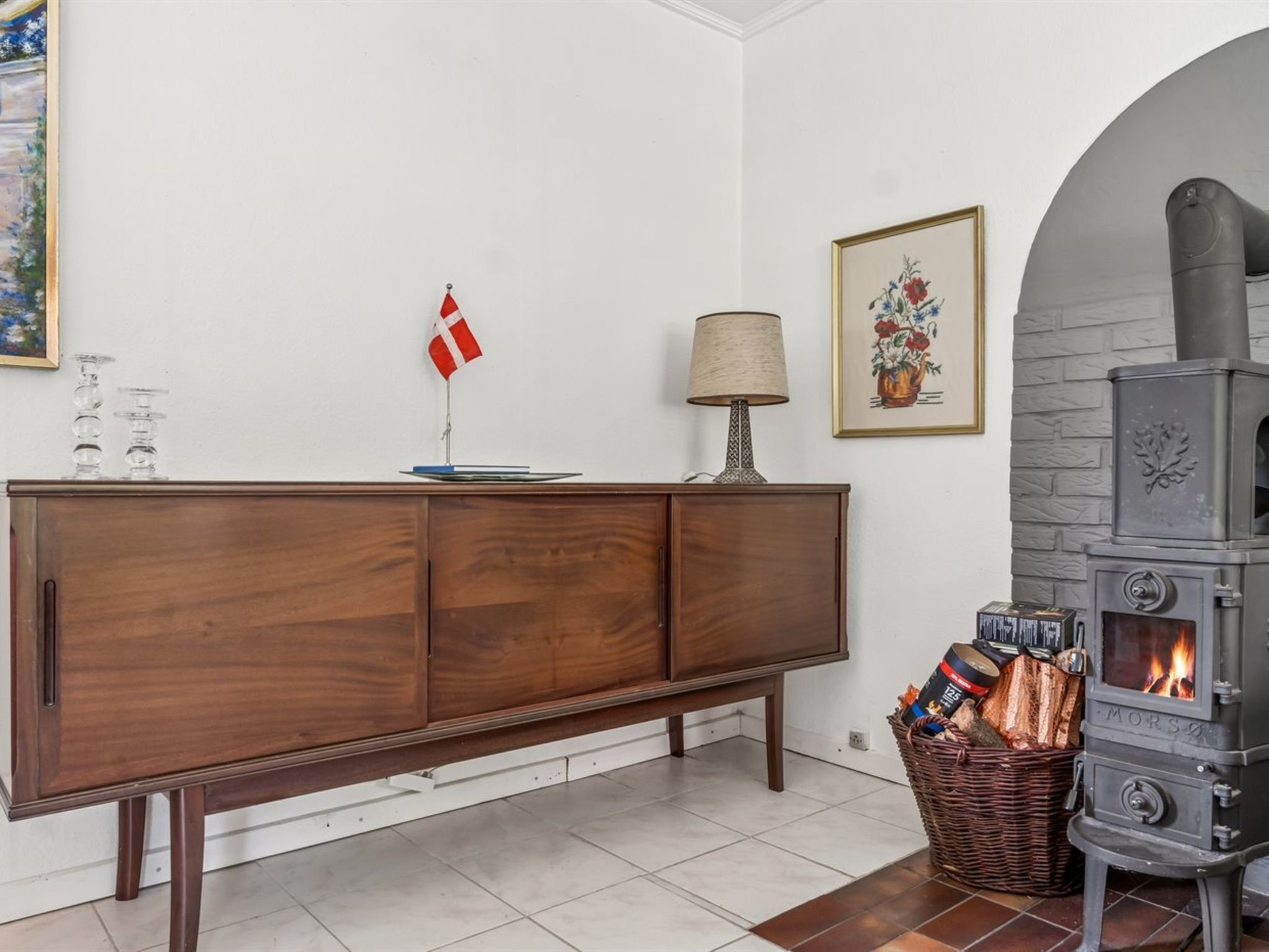 A room with a wooden cabinet, a lamp, a fireplace, and a basket of firewood.