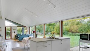 Eine moderne Küche mit großen Fenstern, ein Essbereich mit einem blauen Sofa und einem Fernseher auf einem Schrank.