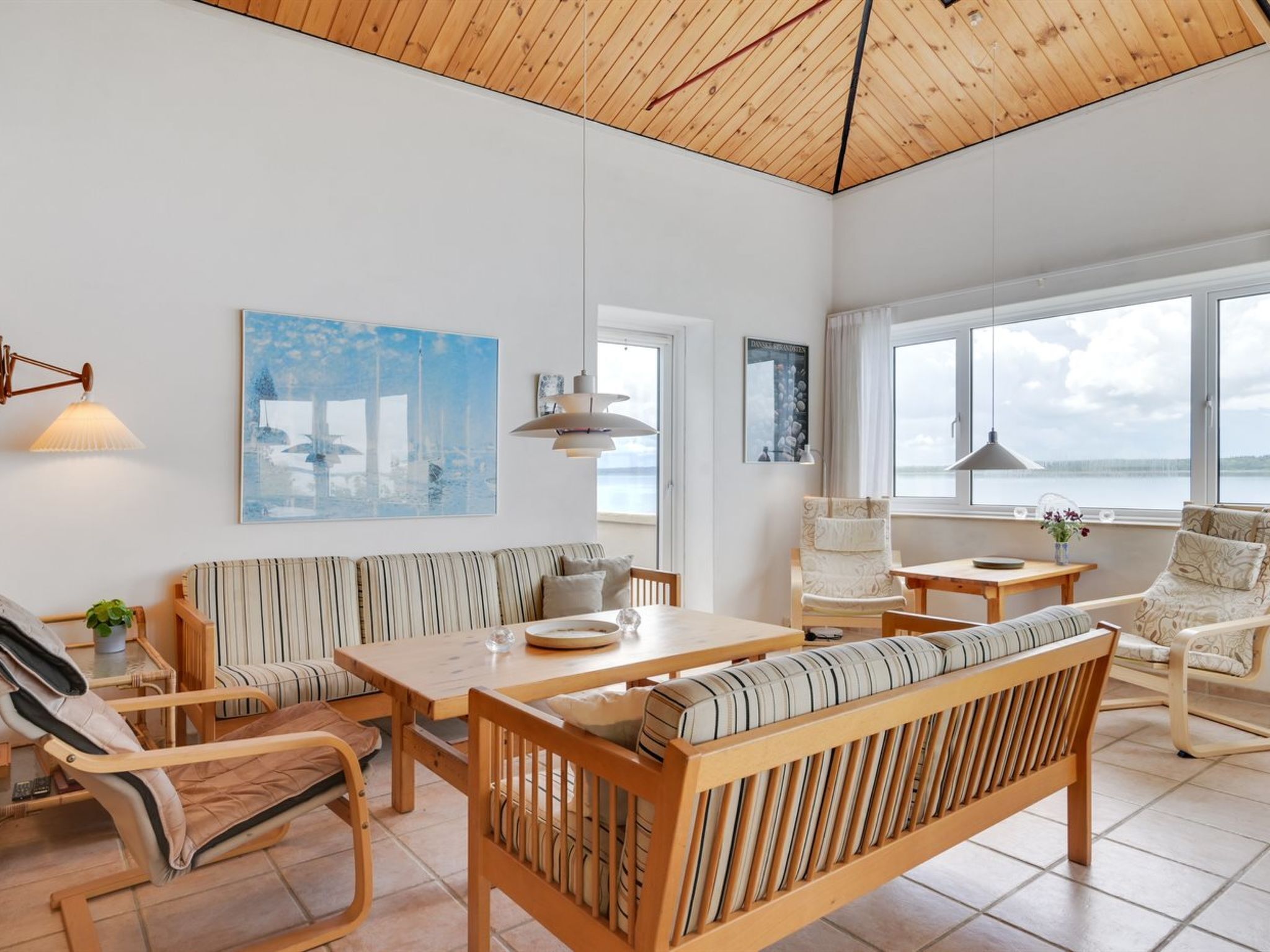 Amplia sala de estar con techo de madera, un ventanal con vista al mar, un sofá a rayas, una mesa de comedor de madera y una silla con cojín.