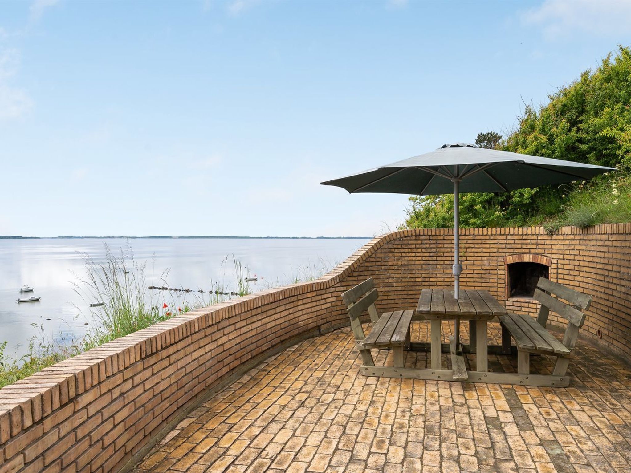 Une terrasse en briques avec une table et un banc en bois, un parasol et une vue sur une étendue d’eau calme.