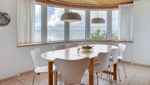 A dining area with a round table, white chairs, and pendant lights. It features large windows with a view of the water and sky. - "Grumme" - 30m to the fjord by Interhome (Ølsted)