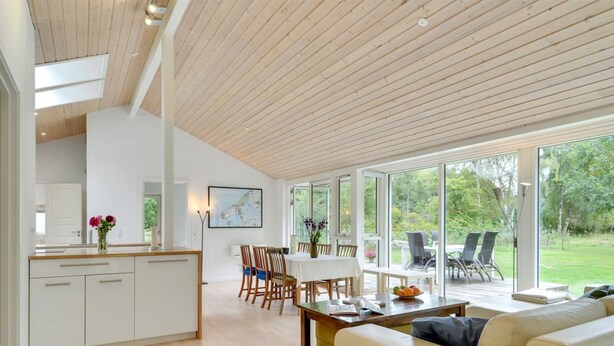 Un vaste espace de vie doté d’un plafond en bois, un coin repas avec une table et des chaises, et de grandes fenêtres offrant une vue sur l’extérieur.