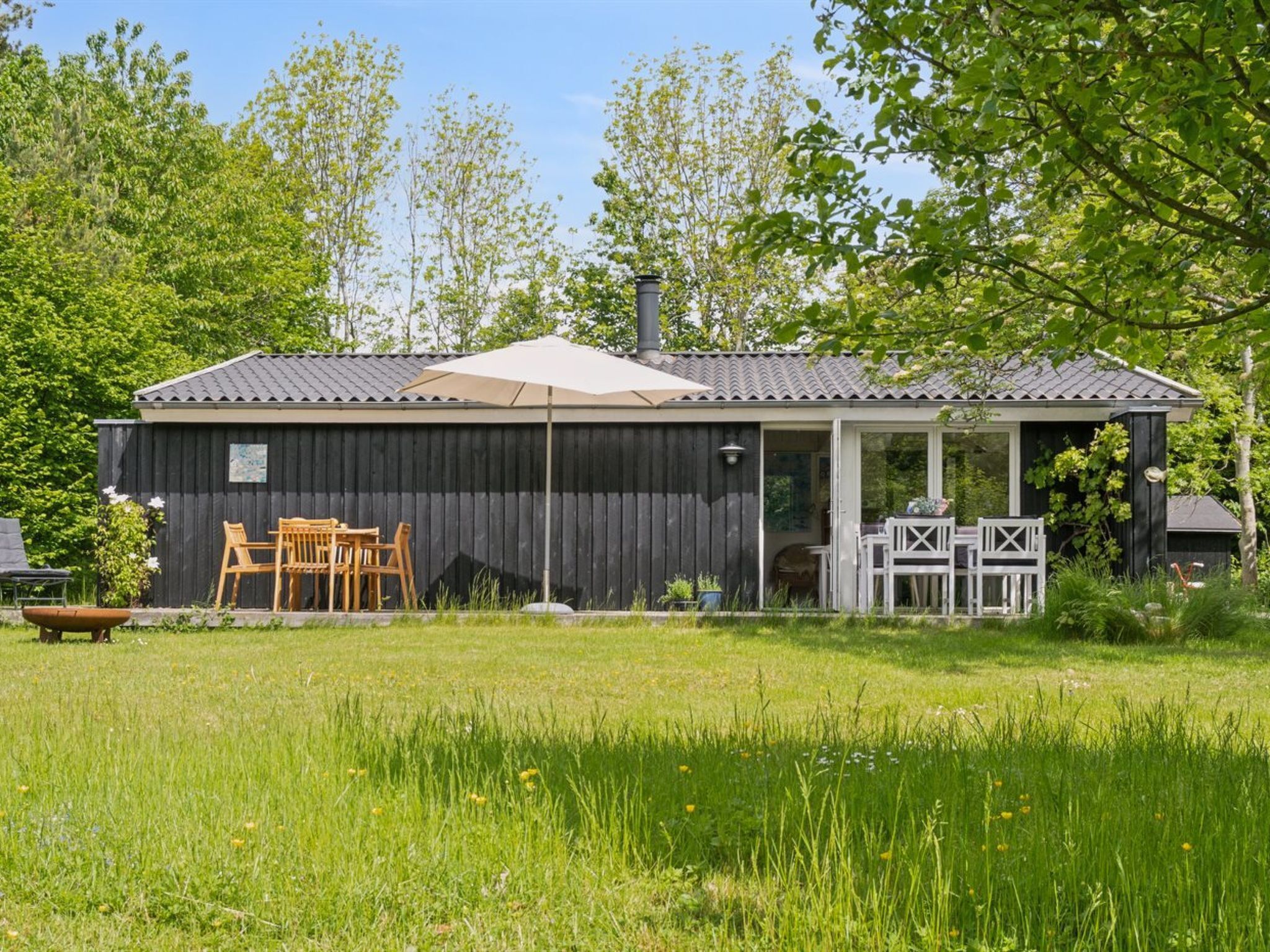 Ein eingeschossiges Gebäude mit dunkler Fassade, einer überdachten Terrasse mit Gartenmöbeln und einem Rasen.