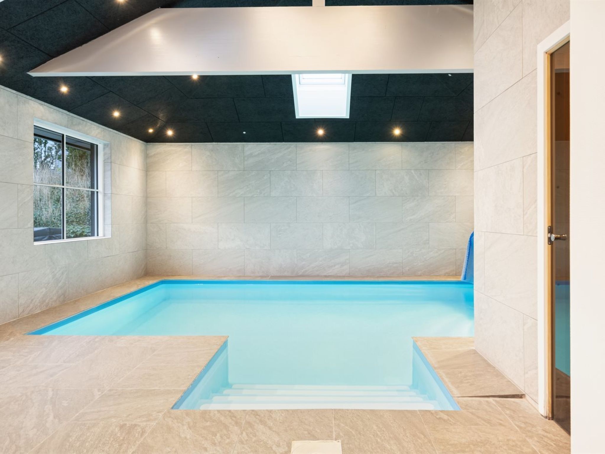 Une piscine intérieure à l’eau d’un bleu limpide, avec des murs carrelés et une fenêtre sur l’un des côtés.