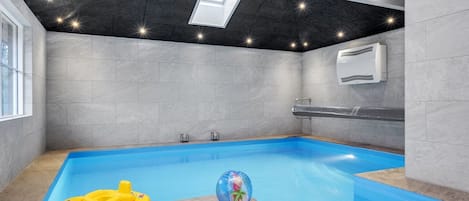 A modern indoor swimming pool with a clear blue water, grey tiled walls, and a skylight.