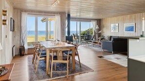 Ein großzügiger Raum mit einem großen Holztisch und Stühlen, einer Couch und einem Fernseher. Große Fenster bieten Blick auf den Strand.