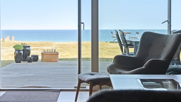 Blick auf einen Strand von einer überdachten Terrasse mit Tisch, Stühlen und Topfpflanzen, bewölkter Himmel.