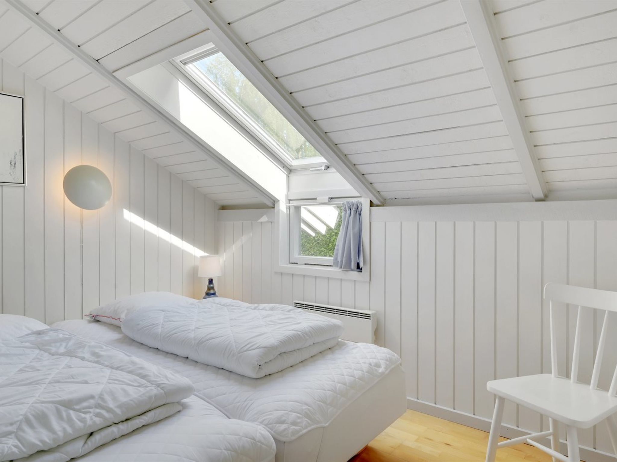 Une chambre avec un plafond en pente, un lit avec une literie blanche, une chaise et une fenêtre avec des rideaux.