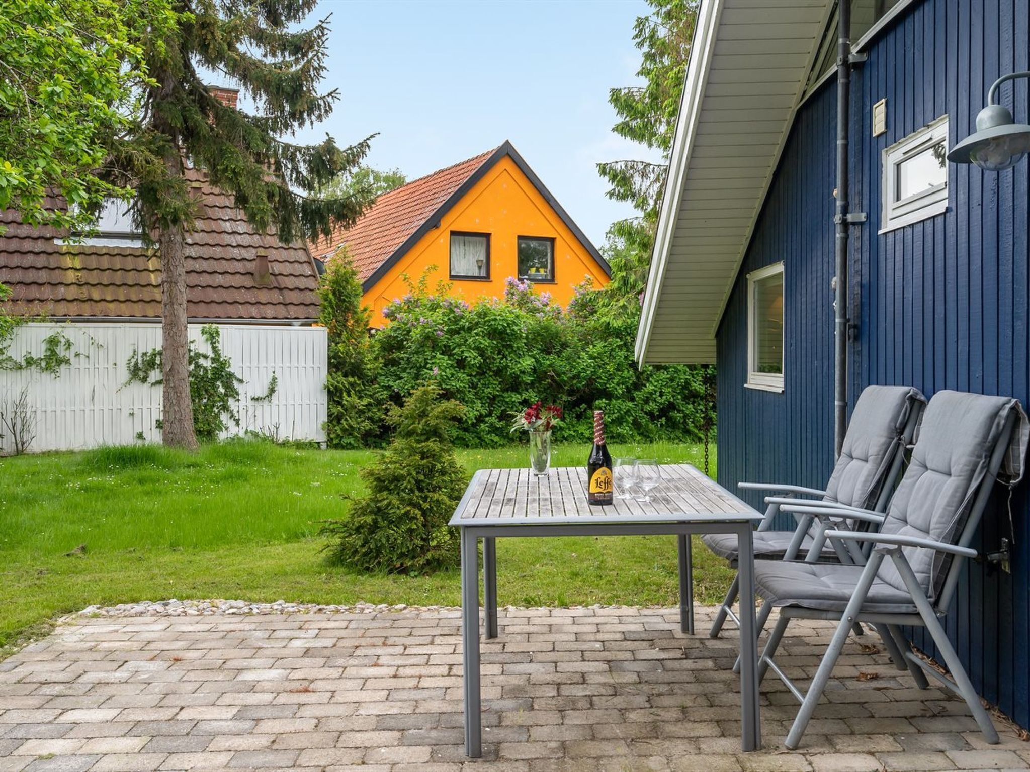 Une terrasse avec une table et des chaises, une bouteille de bière et une vue sur une maison jaune avec un toit rouge.