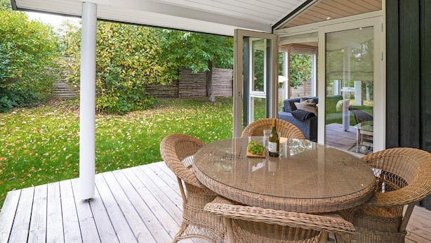 An outdoor patio with wicker furniture and a glass table, surrounded by a wooden deck and a garden.