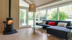 A modern living room with a wood-burning stove, a glass coffee table, a dark sofa, and a view of greenery outside.