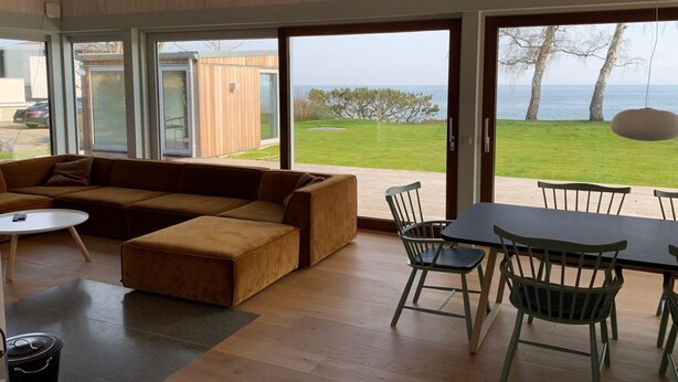 Ein modernes Wohnzimmer mit einer großen Couch, einem Esstisch mit Stühlen und großen Schiebegläsern, die zu einem grünen Außenbereich mit Meerblick führen.