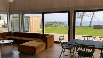 Un salon moderne doté d’un grand canapé, d’une table à manger avec des chaises et de grandes portes-fenêtres coulissantes donnant sur un espace extérieur verdoyant avec vue sur la mer.