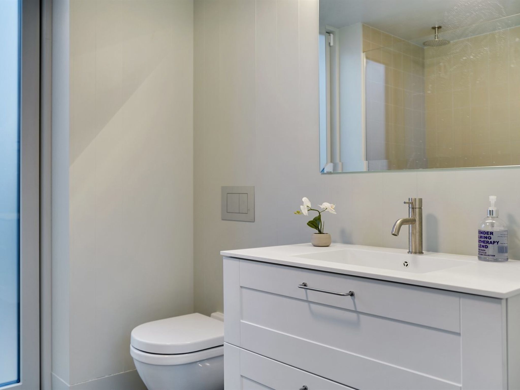 A modern bathroom with a white toilet, a vanity with a mirror, and a bottle of hand sanitizer on the counter.