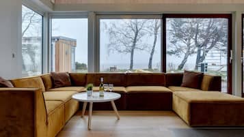 Un salon moderne doté d’un grand canapé d’angle, d’une table basse ronde et offrant une vue sur la mer grâce à de grandes fenêtres.