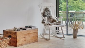 Ett modernt vardagsrum med en vit gungstol, en byrå i trä och utsikt över grönskan genom stora fönster.