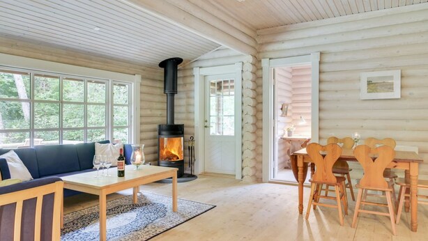 Et interiør af en træhytte med en central pejs, en sofa, et spisebord med stole og store vinduer.