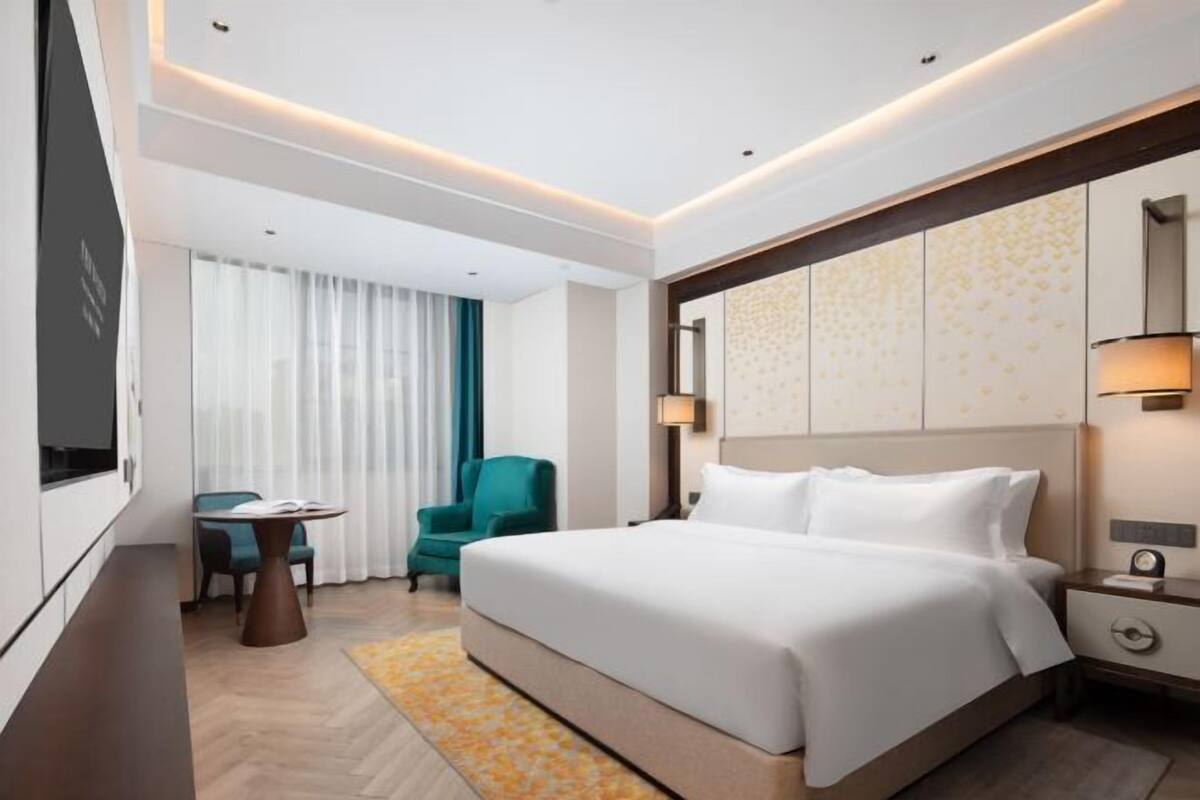 A modern hotel room with a large bed, a desk with a chair, a television, and a wall-mounted lamp.