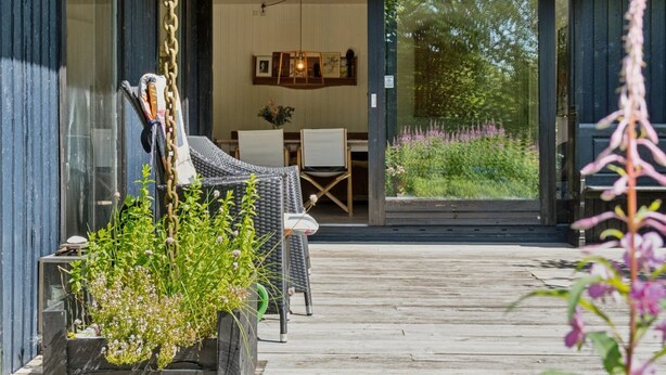 A wooden deck with outdoor seating and a glass door leading to an interior space with a dining table and chairs.