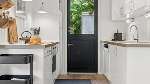 A compact kitchen with white cabinetry, a black door, a clock, and a window with a view of greenery.
