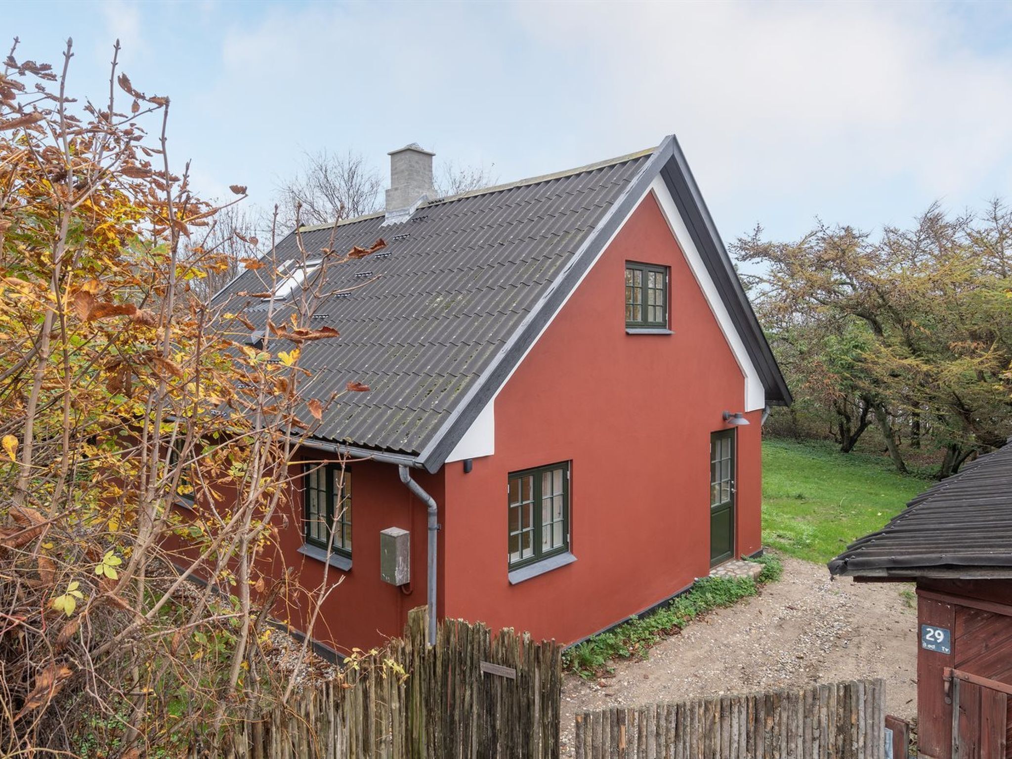 Une maison rouge avec un toit gris, une cheminée et une clôture en bois à l’avant.