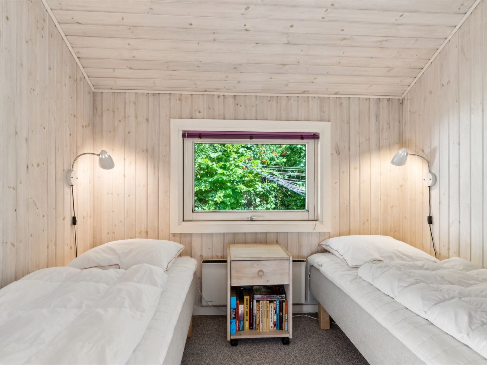 Ein Zimmer mit zwei Betten, einem kleinen Nachttisch mit Büchern, einem Fenster mit Blick auf Grünflächen und Wandlampen.
