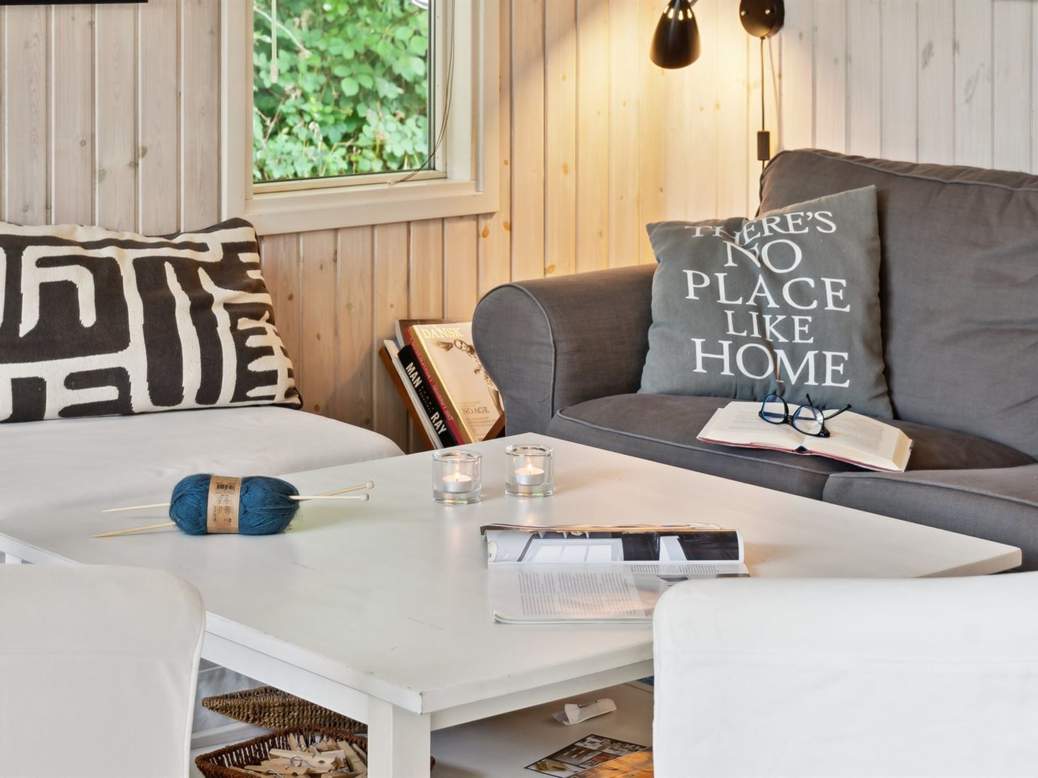 A cozy living room with a white coffee table, grey sofa, and a window with wooden paneled walls.
