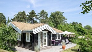 Une cabane en rondins avec un toit de chaume, une terrasse dotée de mobilier d’extérieur et un jardin environnant.