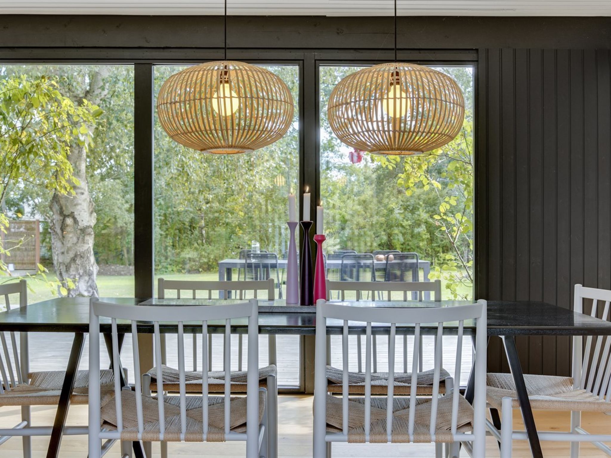 A dining area with a glass table and white chairs, two wicker pendant lights, and a view of a garden with a tree and outdoor furniture.